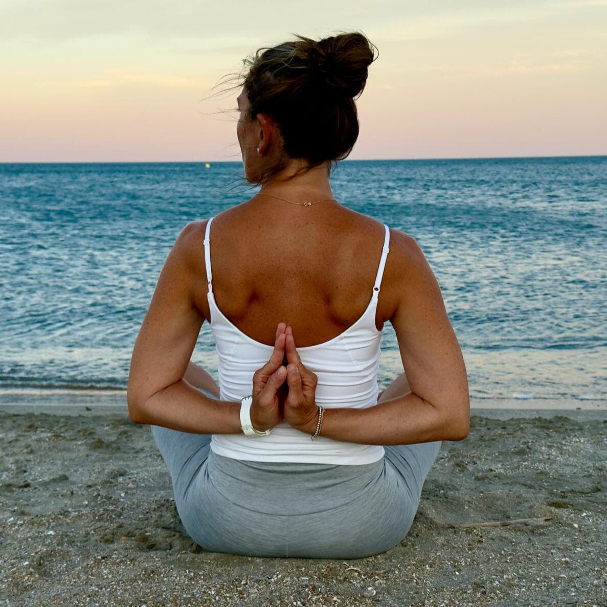 Nina sitzt im Meditationssitz am Strand, die Hände hinter dem Rücken gefaltet, mit Blick aufs Meer